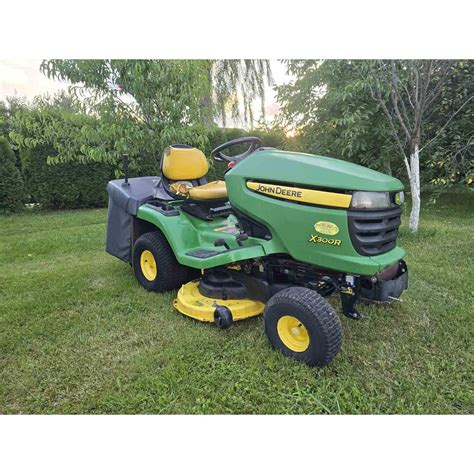 Vizualizacija: John Deere traktoriukas su pilnu 300 litrų žolės bunkeriu, rodomas žolės išpylimo mechanizmas.